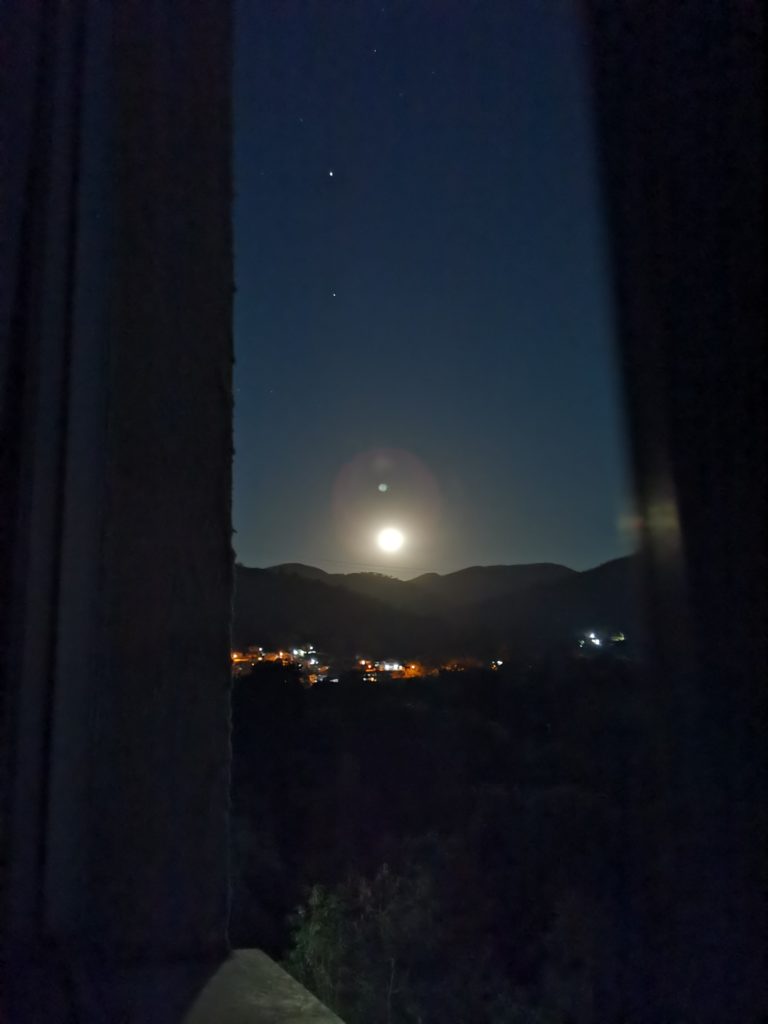 paisagem com luzes de cidade, montanhas na linha do horizonte e a lua brilhando logo acima dos montes